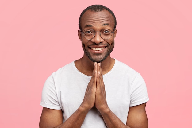 Man with broad shining smile, keeps hands in praying gesture, asks for something desirable