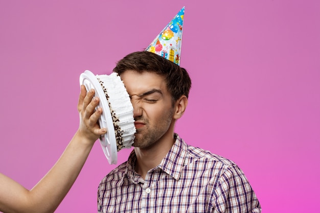 Free photo man with cake on face over purple wall. birthday party.