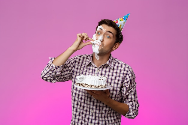 Man with cake on face over purple wall. Birthday party.