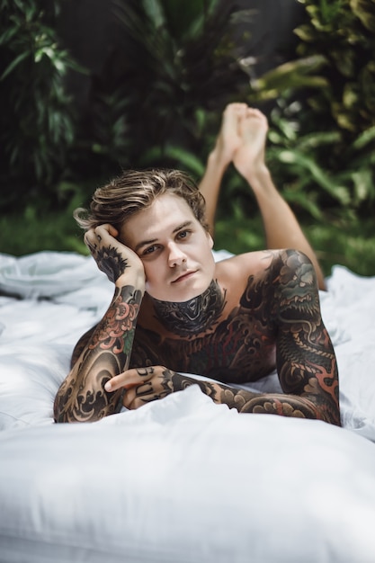 Free Photo man with colorful tattoos resting on a white sheet in the garden 