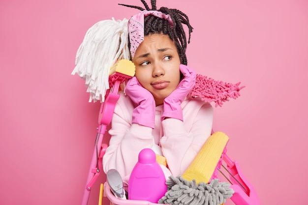 Free photo man with dreadlocks holds chin surrounded by cleaning supplies holds near basin of laundry isolated on pink