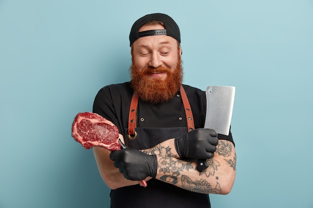 Free photo man with ginger beard in apron and gloves holding knife and meat