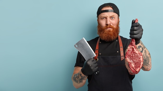 Free photo man with ginger beard in apron and gloves holding knife and meat