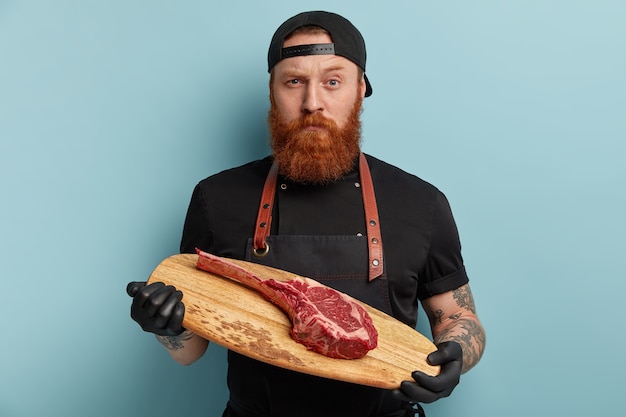 Free Photo man with ginger beard in apron and gloves holding wooden board with piece of meat