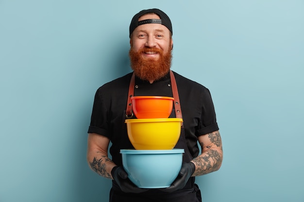 Man with ginger beard wearing apron and gloves holding pile of bowls