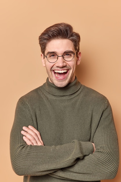 Free Photo man with glad face expression keeps arms folded has positive talk with friend discuss something funny wears warm jumper poses on beige.