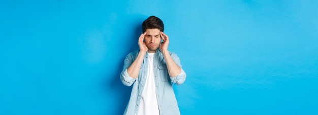 Free Photo man with hangover touching head and grimacing having headache standing against blue background