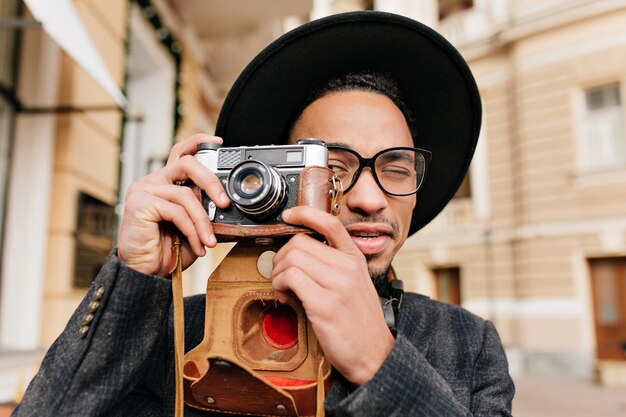 Man with light-brown skin taking pictures with camera. Outdoor close-up portrait of black male photographer wears hat in cold day.