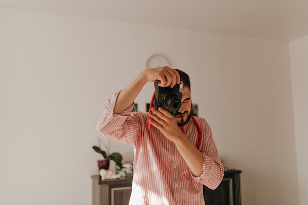 Free photo man with ring on his finger makes photo on camera. snapshot of brunet guy in striped outfit in spacious apartment.