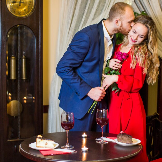 Man with roses kissing pretty woman on cheek in restaurant