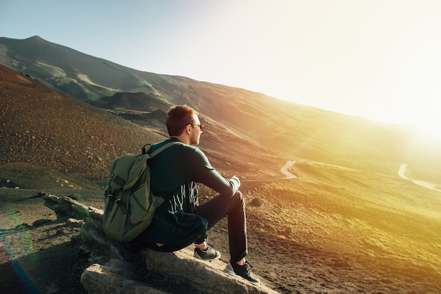 Man with rucksack sitting on rock at sunset on volcano Etna mountain in Sicily