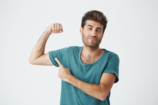 Free photo man with stylish hairstyle and beard frowning his eyebrow, demonstrating how strong he is. fit guy pointing with his finger at his biceps. macho man showing himself