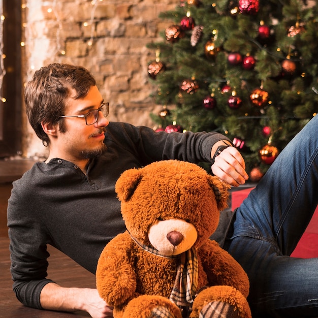 Free photo man with teddy on floor in front of christmas tree