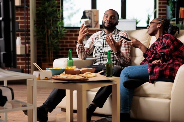 Man and woman enjoying fast food delivery and watching tv, switching channels to find movie on television. Eating takeaway meal package and drinking beer from bottle, leisure activity.