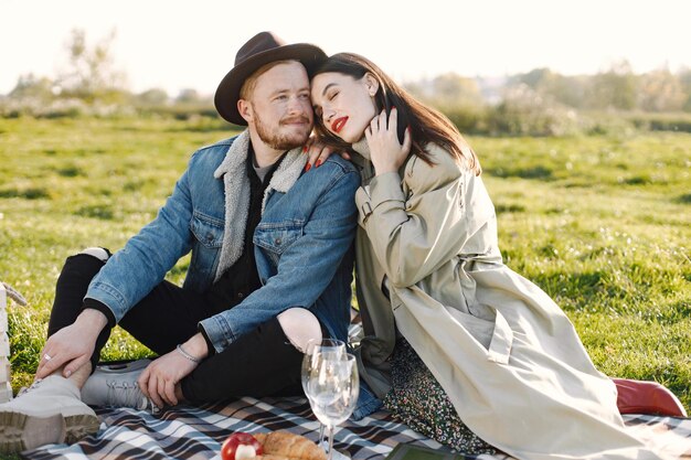 Man and woman in fashion clothes sitting on a nature on a picnic rug. Man wearing jacket and a black hat and woman coat