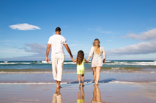 Man, woman and kid wearing pale summer clothes, walking on wet sand to sea, spending leisure time on beach