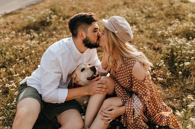 Free photo man and woman kiss in park. couple enjoying great day sitting on grass with their labrador.