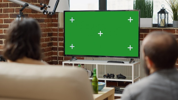 Free photo man and woman watching green screen on television display in living room. people looking at isolated copy space with blank mockup template and chroma key background on tv. tripod shot. close up.