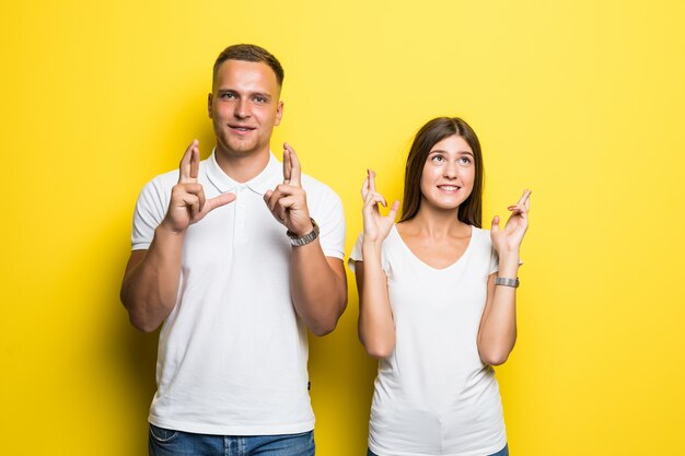 Man and woman in white t-shirts holding fingers crossed isolated on yellow background