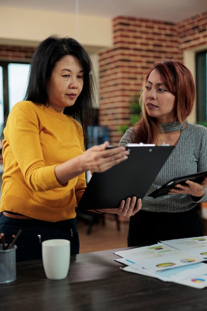 Manager standing beside entrepreneur looking at papers with company graphs analyzing statistics working at management strategy during business meeting in startup office. Modern workplace