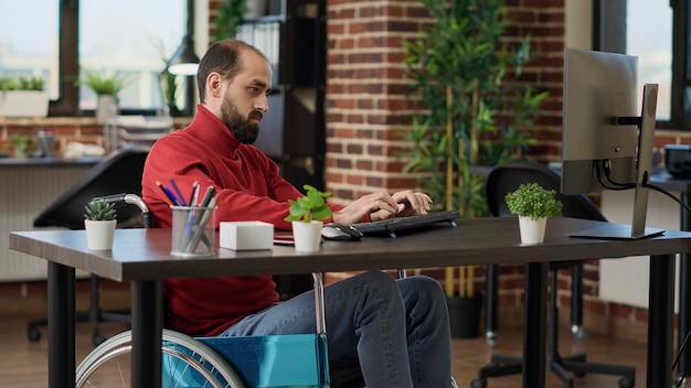 Manager with impairment working on business growth with computer at desk, witting in wheelchair. Male worker planning financial strategy to create executive company development.