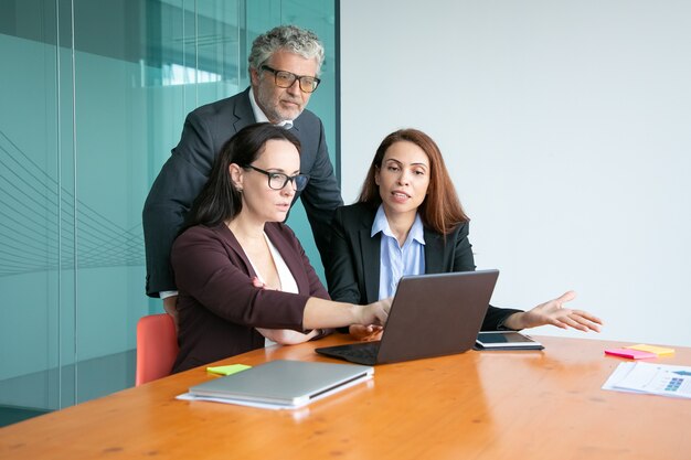 Managers showing presentation on laptop to executive, pointing at display, explaining details.