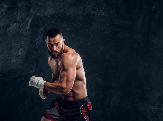 Manly bearded man with beautiful muscular torso is posing for photographer on the dark background.