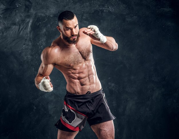 Manly bearded man with beautiful muscular torso is posing for photographer on the dark background.