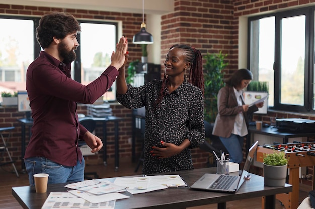 Marketing company employees congratulating each other for great startup marketing strategy results. Happy smiling pregnant woman high fiving coworker for amazing presentation ideas.