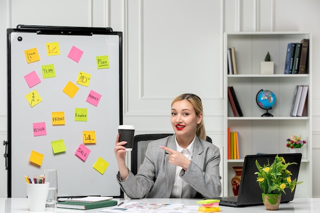 Free photo marketing young cute blonde girl in grey suit in the office pointing at paper cup