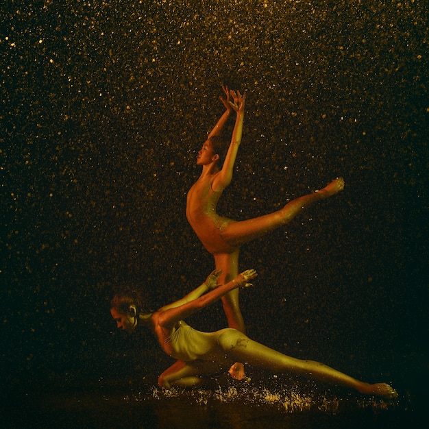 Marvelous. Two young female ballet dancers under water drops and spray. Caucasian and asian models dancing together in neon lights. Ballet and contemporary choreography concept. Creative art photo.