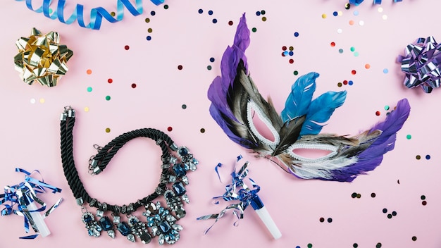 Masquerade carnival feather mask with confetti and necklace over pink backdrop