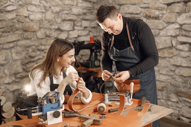 Free Photo mature craftsman with young woman working in workspace man and woman wearing an apron and making a belt grounge dark stone texture background