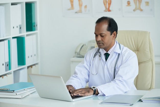 Mature Doctor Sitting At Desk Working On Laptop