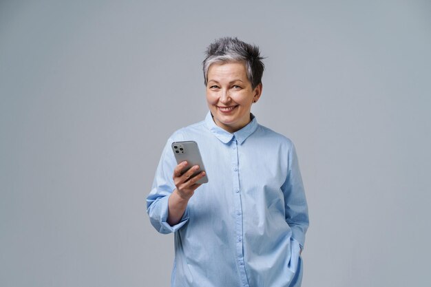 Mature grey haired businesswoman with smartphone in hand looking at camera shopping online checking social media Pretty woman in blue shirt isolated on white background Copy space