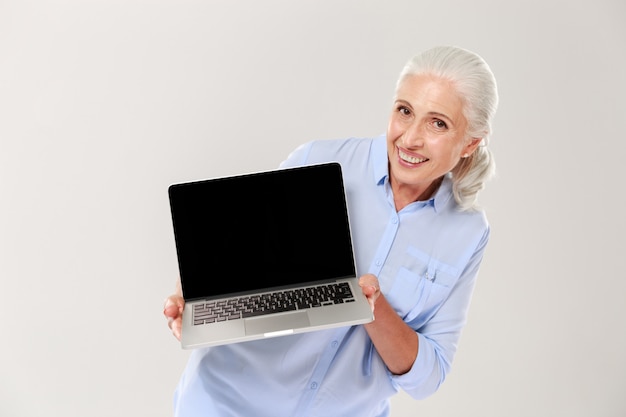 Mature grey-haired woman smiling and showing blank screen of laptop isolated