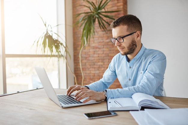 Mature male freelance web designer sitting in co-working space, working on laptop computer, writing down tasks in notebook