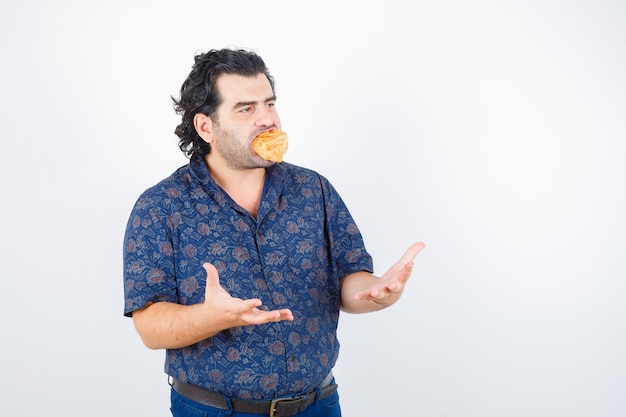 Mature man holding pastry product on mouth while keeping hands in aggressive manner in shirt and looking serious , front view.