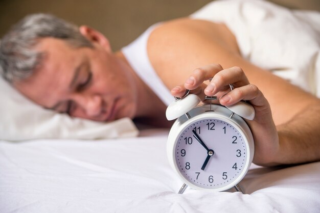 Mature man sleeping in bed with alarm clock in foreground at bedroom. Exhausted man being awakened by an alarm clock in his bedroom. Waked Up