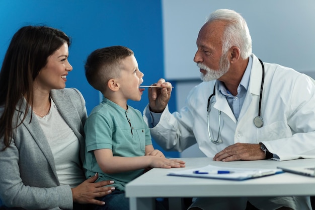 Mature pediatrician examining throat of a little boy who came to medical appointment with his mother