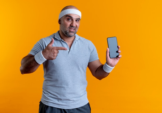 Mature sporty man in headband showing smartphone pointing with finger to it looking confident standing over orange wall