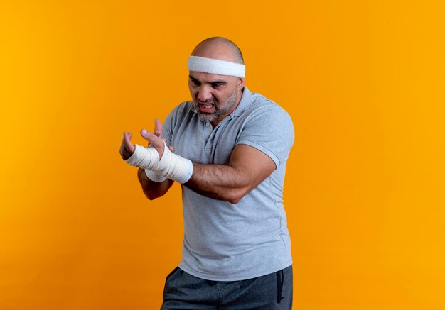 Mature sporty man in headband touching his bandaged hand looking unwell suffering from pain standing over orange wall