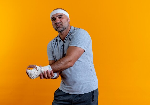 Mature sporty man in headband touching his hand looking unwell having pain standing over orange wall