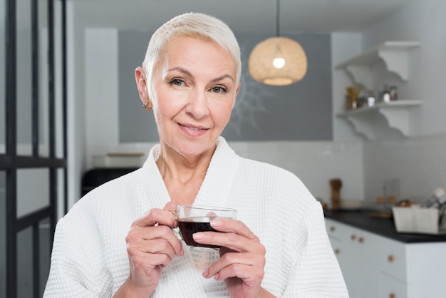 Free photo mature woman in bathrobe posing in the kitchen while holding coffee cup