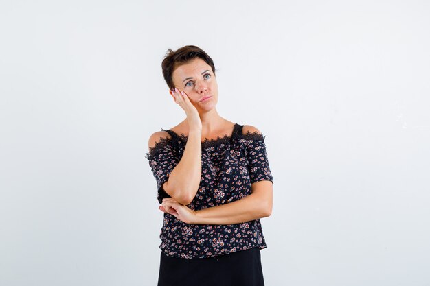 Mature woman in floral blouse, black skirt leaning cheek on palm, thinking about something and looking pensive , front view.