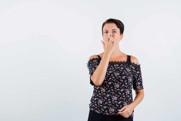 Mature woman in floral blouse and black skirt pulling nose upward with fingers and looking serious , front view.