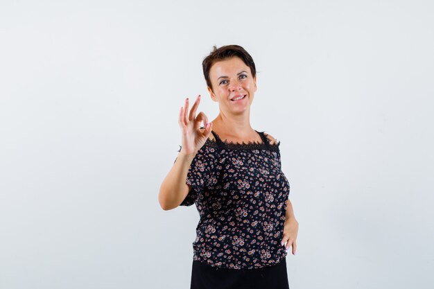 Mature woman in floral blouse and black skirt showing ok sign and looking cheerful , front view.