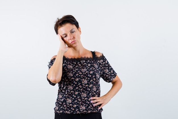 Mature woman holding hand on waist, leaning cheek on palm, looking down in floral blouse, black skirt and looking pensive. front view.