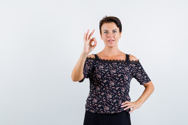 Mature woman holding one hand on waist, showing ok sign in floral blouse and black skirt and looking confident. front view.