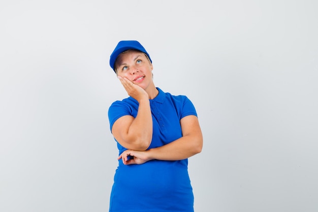 Mature woman leaning cheek on raised palm in blue t-shirt and looking hopeful.